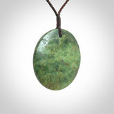 This piece is a large sized, oval round, disc pendant. It is suspended on a brown coloured braided cord that is length adjustable.